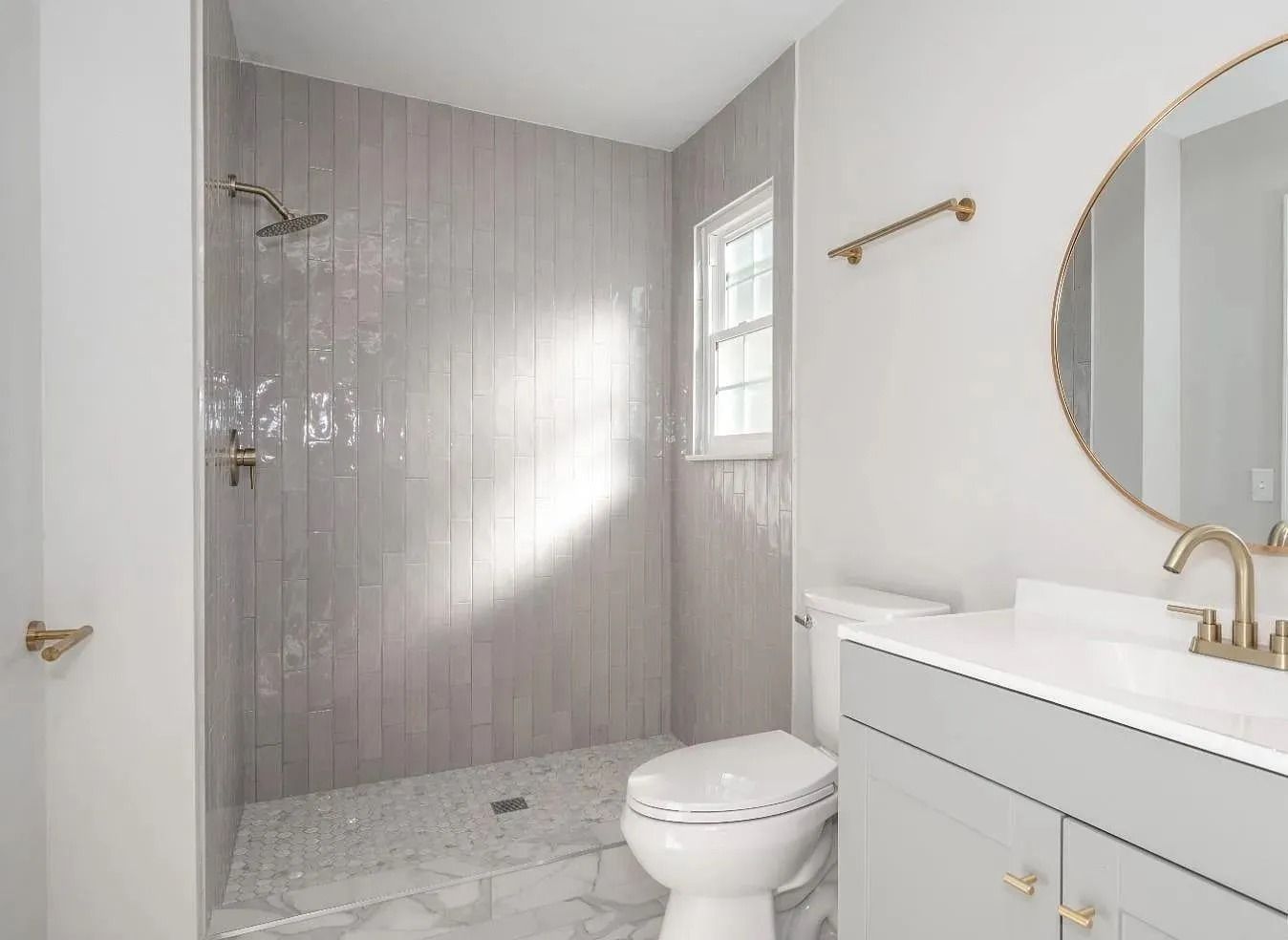Modern bathroom with gray tile shower, white toilet and vanity, gold fixtures, and round mirror.