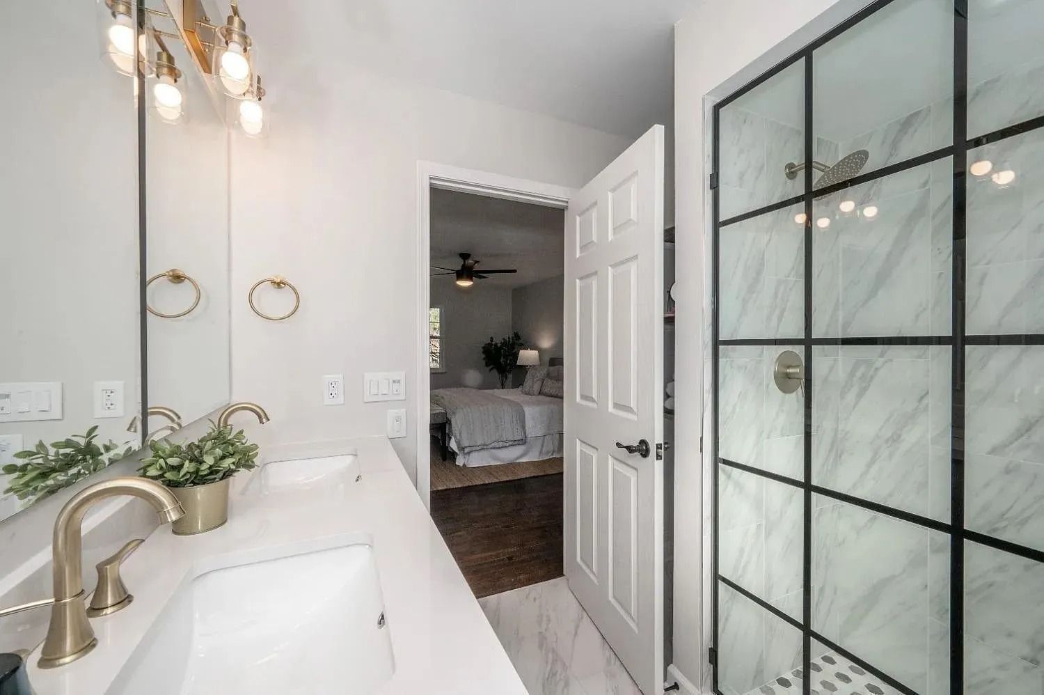 Modern bathroom with white countertops, gold fixtures, glass shower, and a doorway to a bedroom.