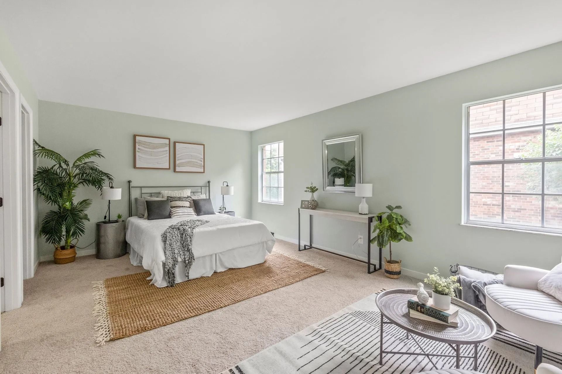 Bedroom with bed, rug, plants, and seating in front of a window. Light green walls and light-colored carpet.