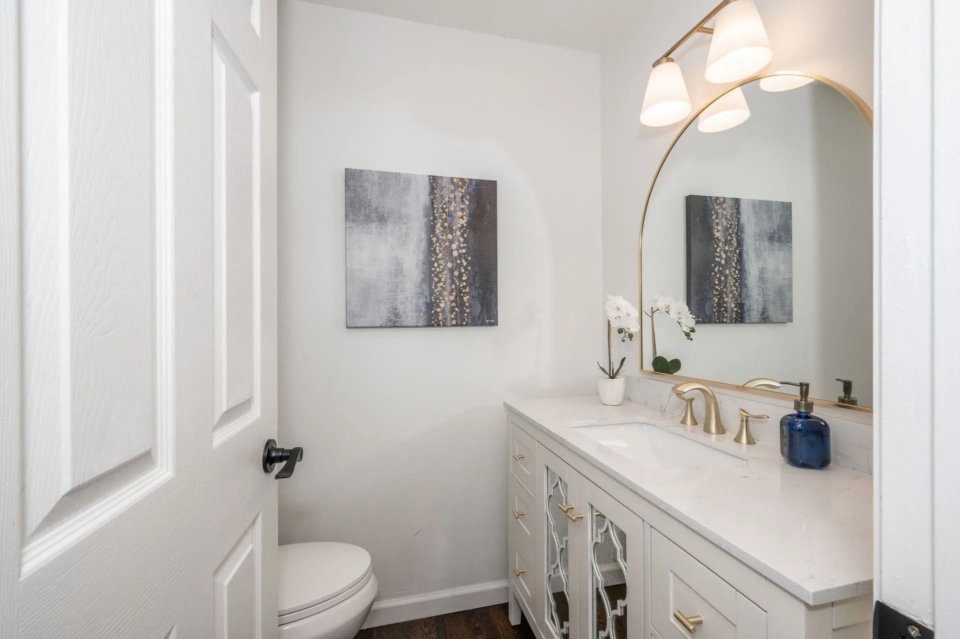 A small white bathroom with a vanity, toilet, and artwork. Gold accents and a decorative mirror are featured.