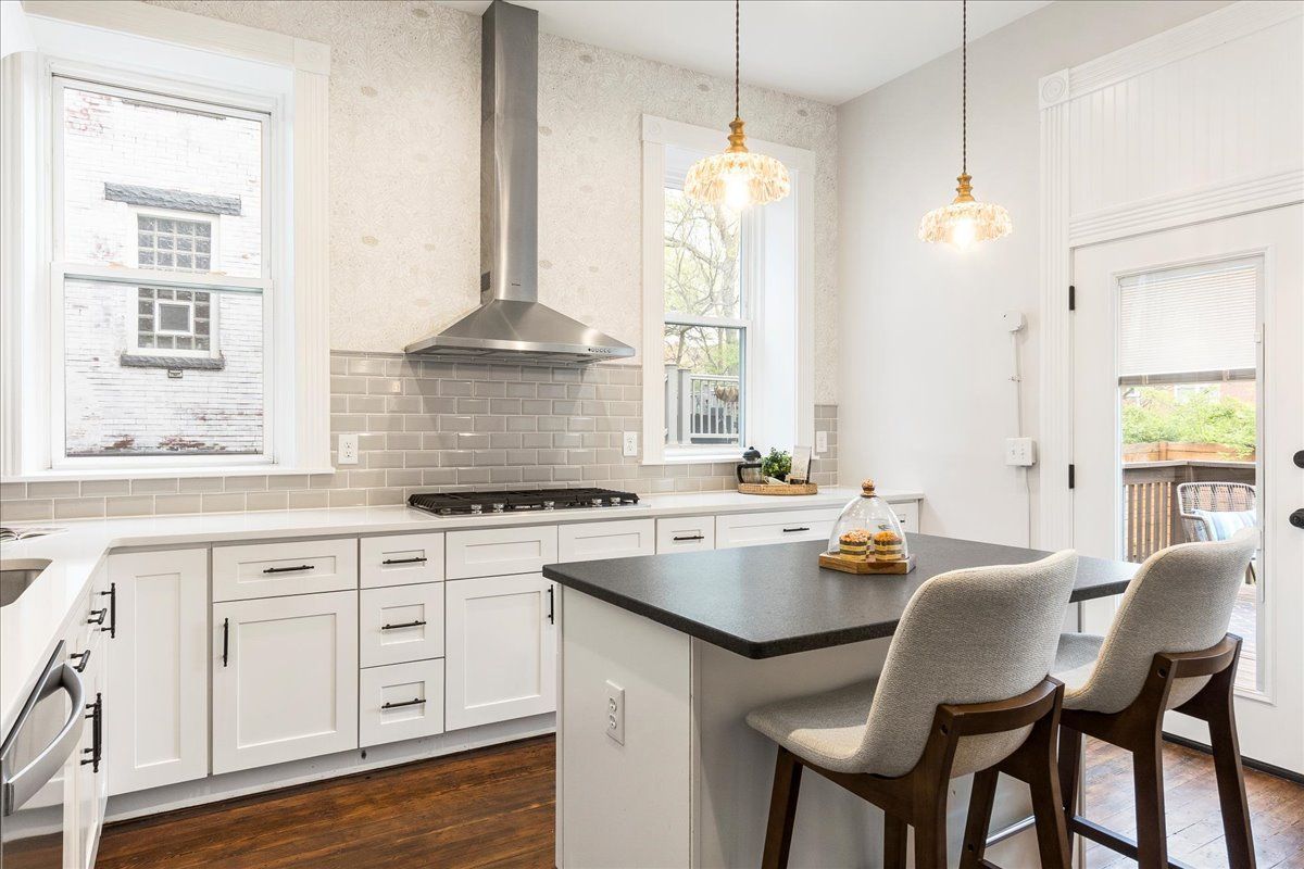 Kitchen with white cabinets, stainless steel range hood, island with two chairs, and two pendant lights.