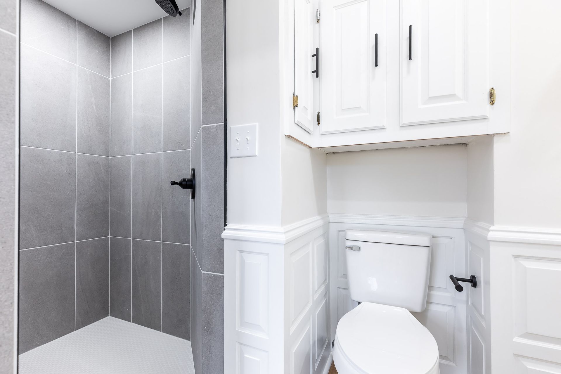 Modern bathroom with gray tiled shower and white toilet below a cabinet.