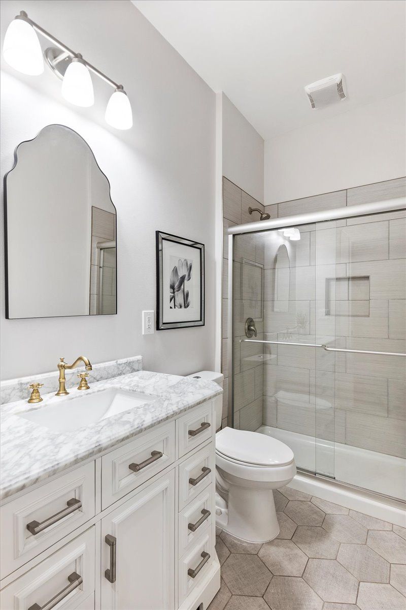 White bathroom with marble vanity, arched mirror, glass shower, and hexagonal floor tiles.