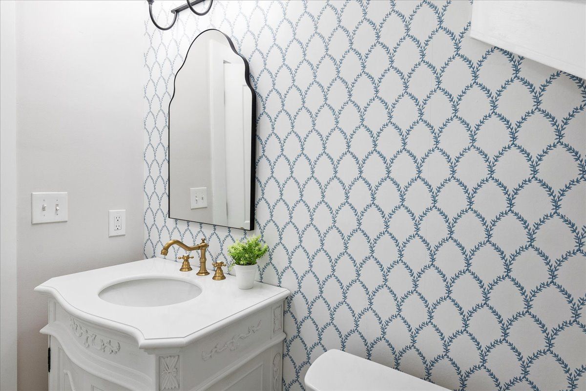 Bathroom with white vanity, blue and white patterned wallpaper, gold faucet, and black-framed mirror.
