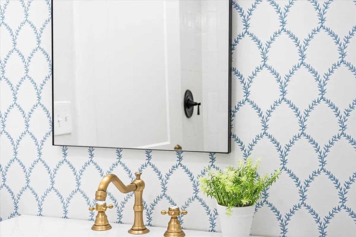 Bathroom with white floral tile, gold faucet, black mirror, and green plant.