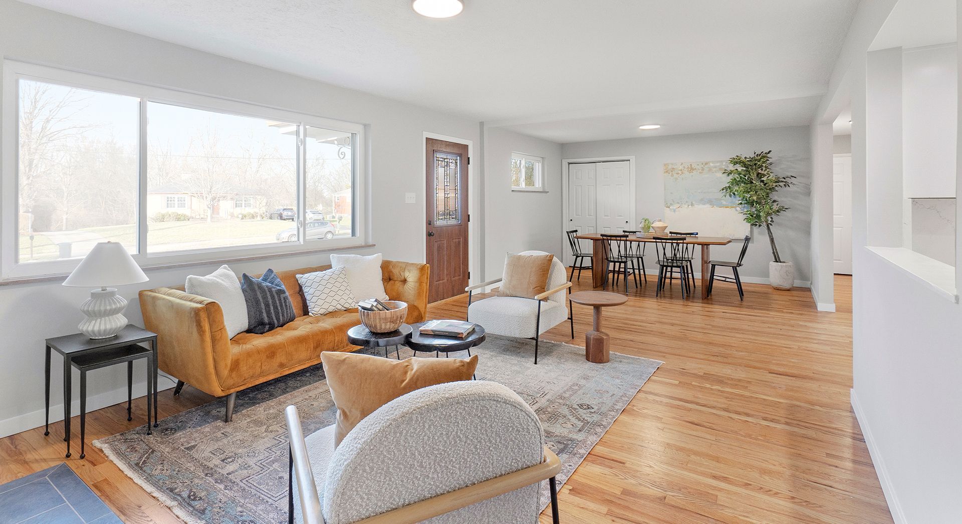 Living room with mustard-colored sofa, rug, and dining area. Large window and hardwood floors.