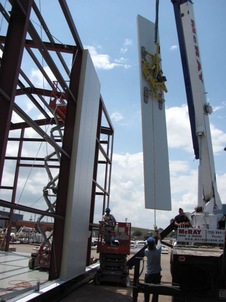 A crane lifting a large white wall panel to be installed on a steel-framed building structure. Workers supervise.