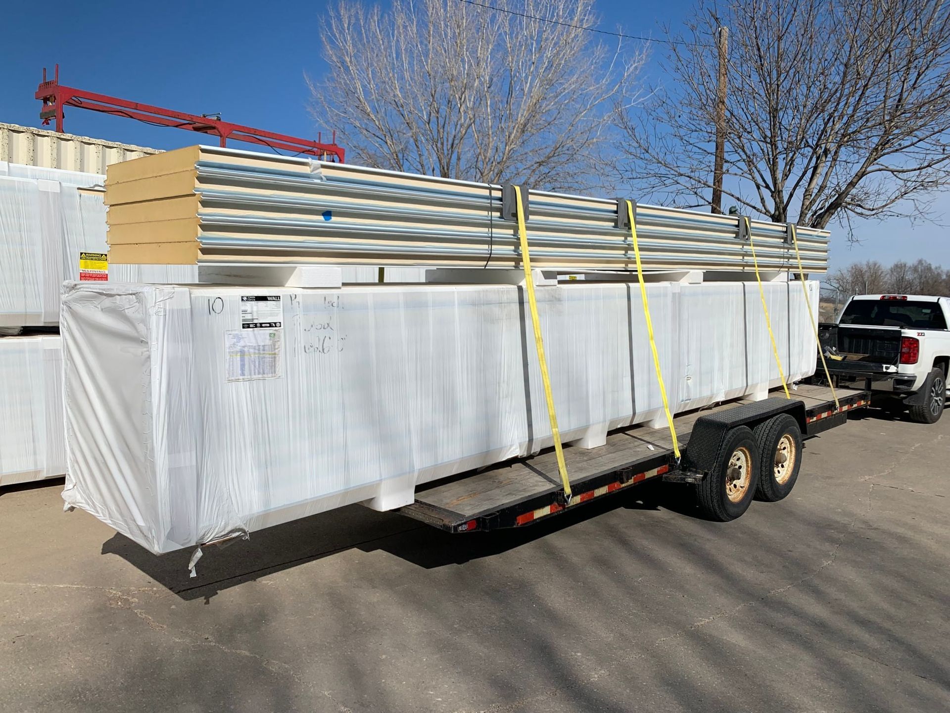 Materials wrapped in white plastic strapped onto a trailer, pulled by a pickup truck. Sunny day.