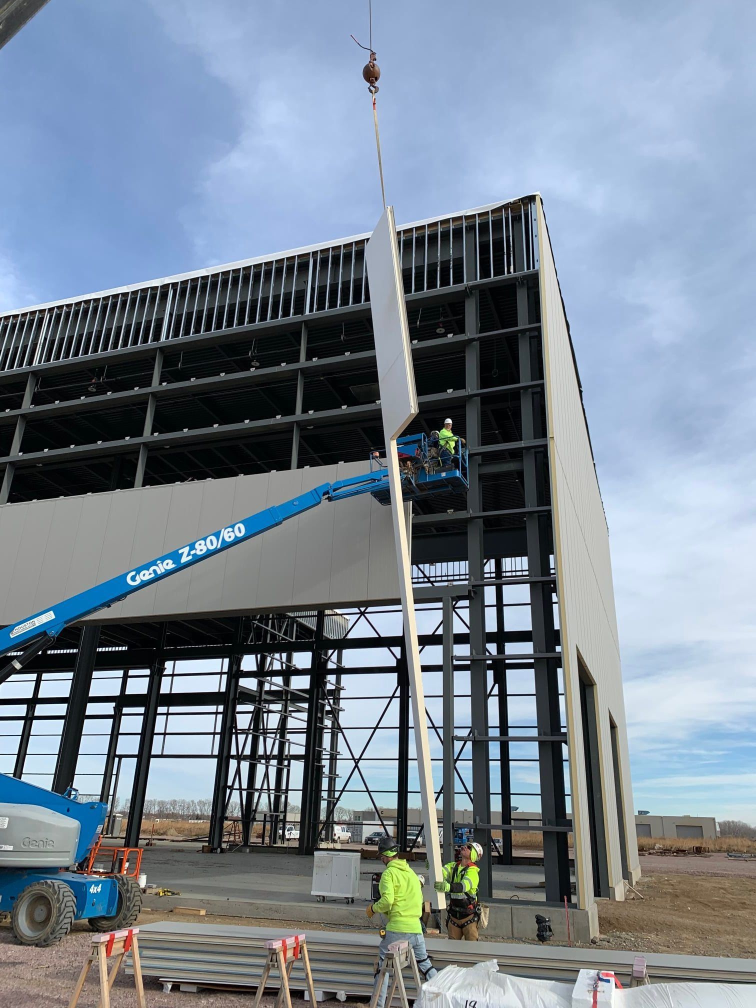 Construction workers installing a wall panel on a steel-framed building, using a crane and lift.