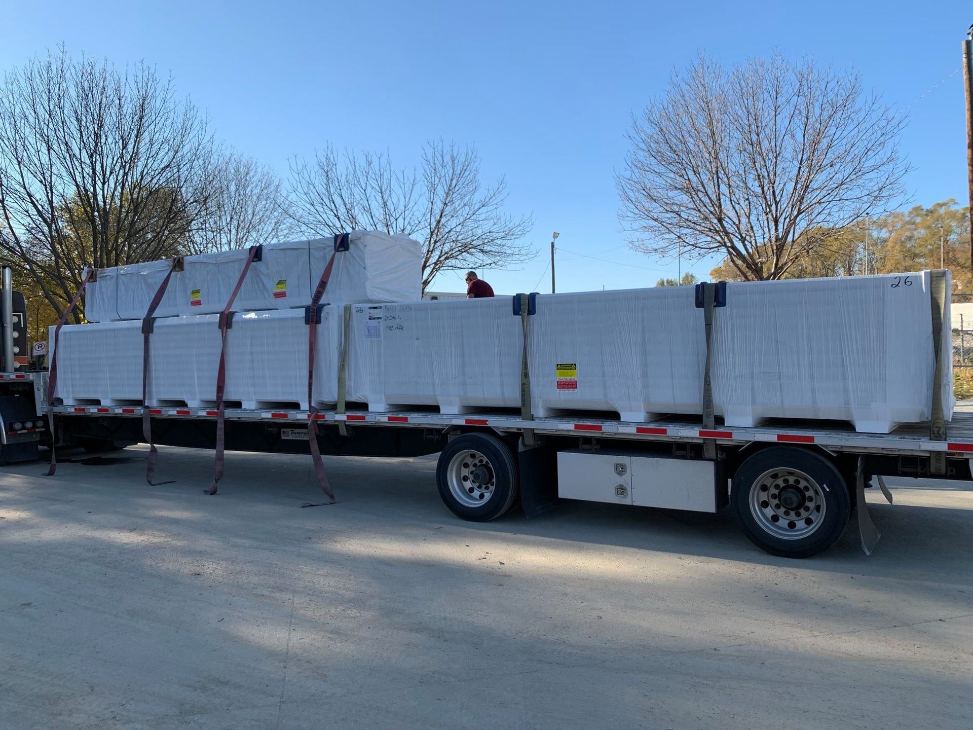 Truck carrying multiple pallets of wrapped building materials on a gravel road, under a blue sky.