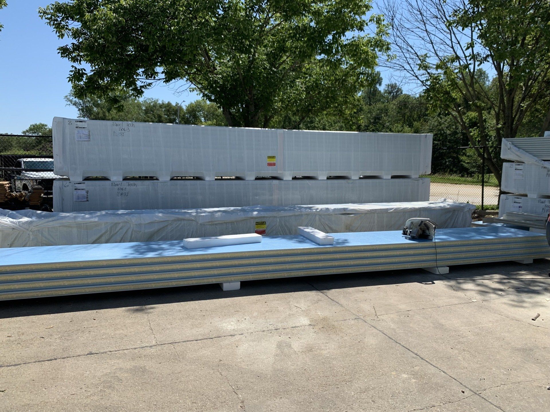 Stacks of white and blue insulated building panels on a concrete surface, outdoors on a sunny day.