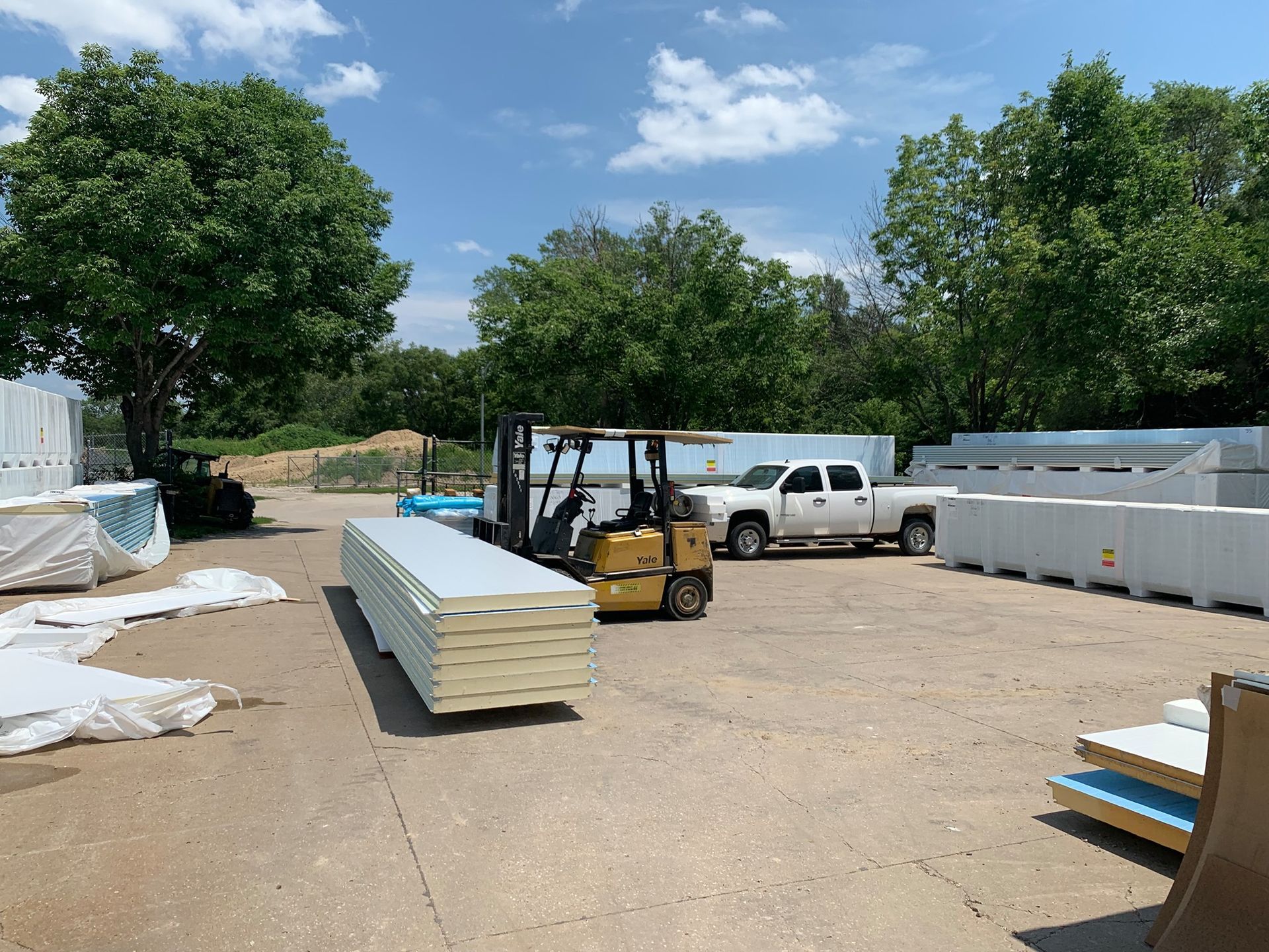 Forklift moving stacked insulated panels near a truck and trees on a sunny day.