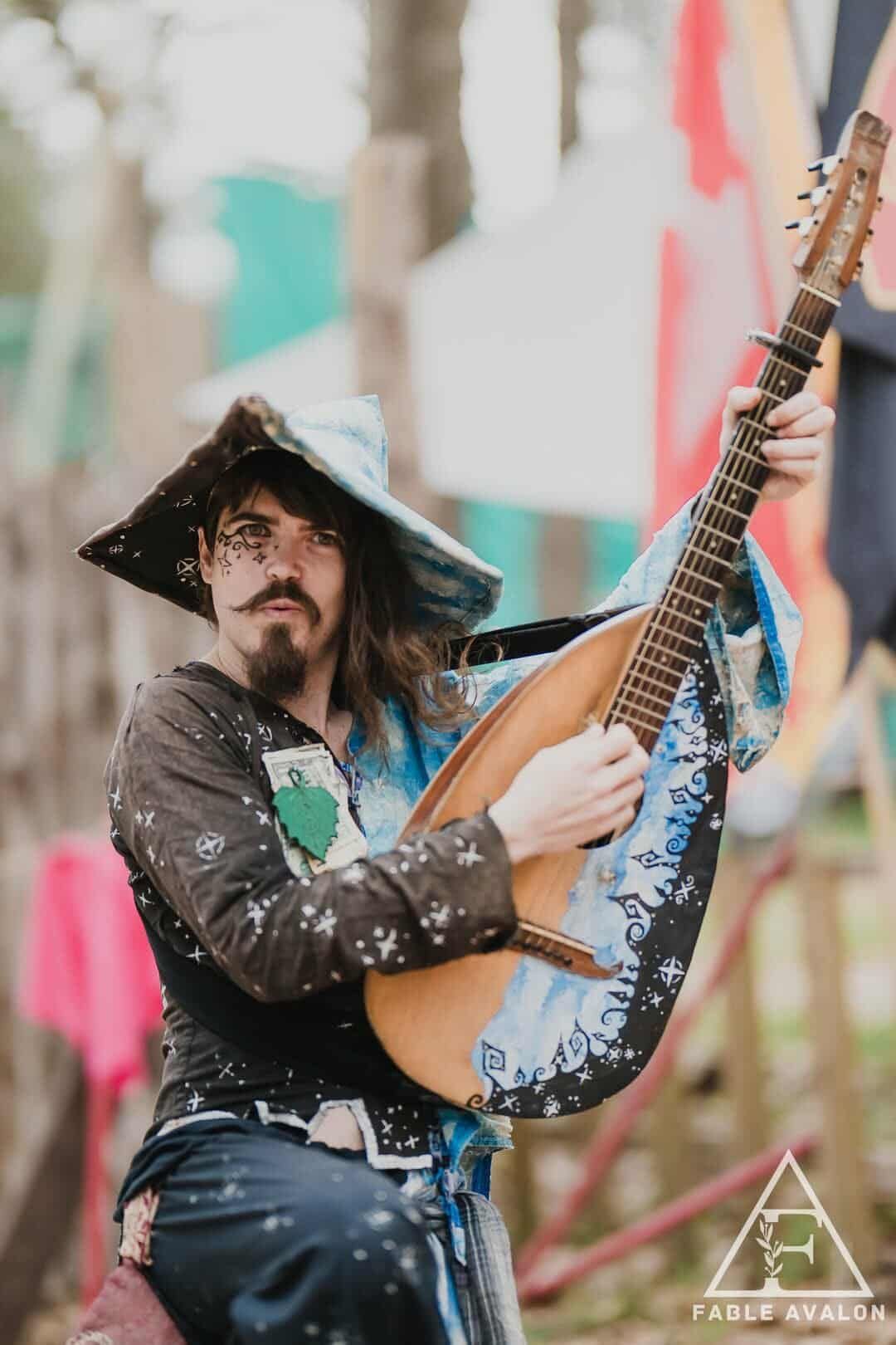 A person in fantasy attire plays a lute outdoors. They wear a hat and look at the instrument, smiling.