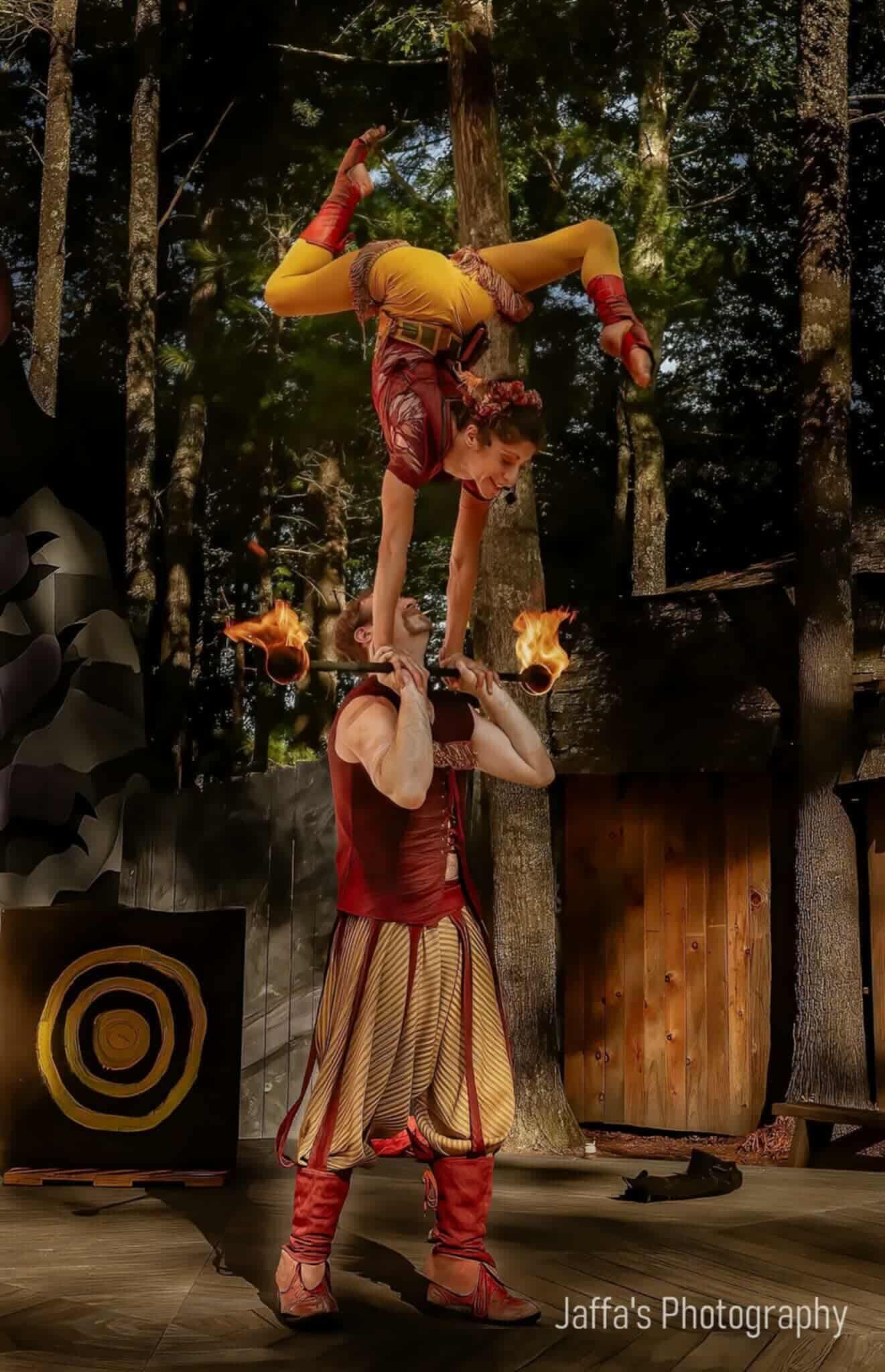Acrobats in Renaissance attire balance, one on top with legs overhead, torches lit in hands. Outdoors.