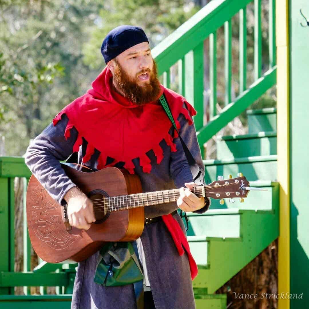 Man in medieval clothing playing acoustic guitar on a wooden stage; red cowl, blue beret.