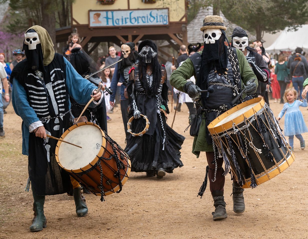 People in costumes dancing at a festival. Many wear dark robes and skull masks. Outdoor setting.