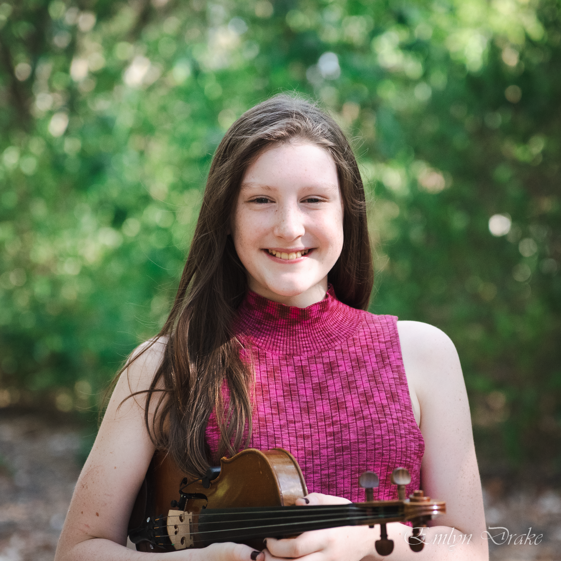 A photo of Emlyn Drake posing with a violin in front of a cozy-looking fireplace.