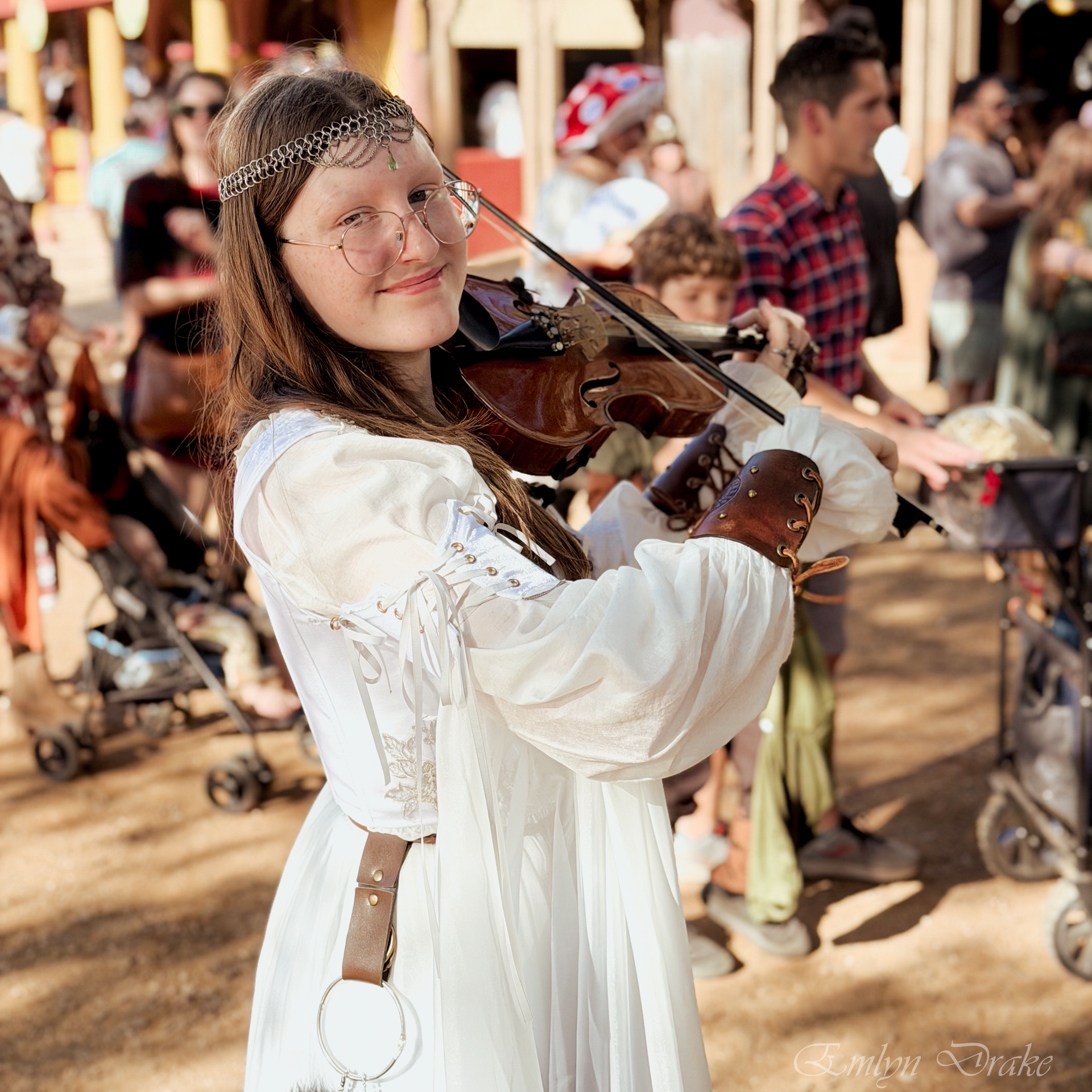 A photo of Emlyn Drake posing with a violin in front of a cozy-looking fireplace.