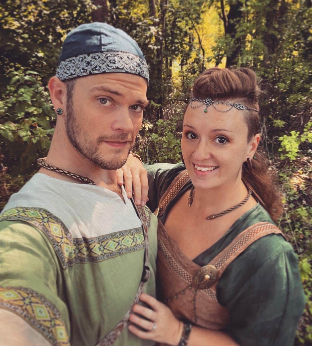 Couple in Renaissance festival attire, smiling and holding hands, with mugs in hand. Outdoors.