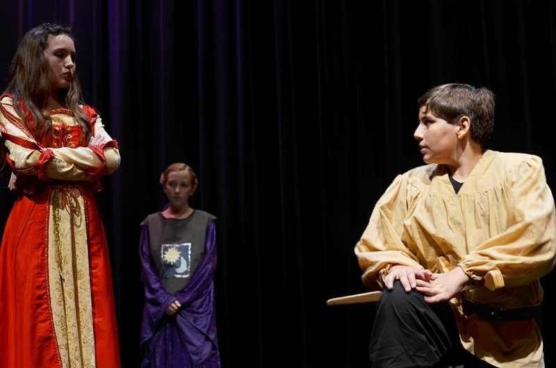 Three actors on a stage. One in red dress, arms crossed. Another in purple robe. Last one in yellow tunic, kneeling. Black backdrop.