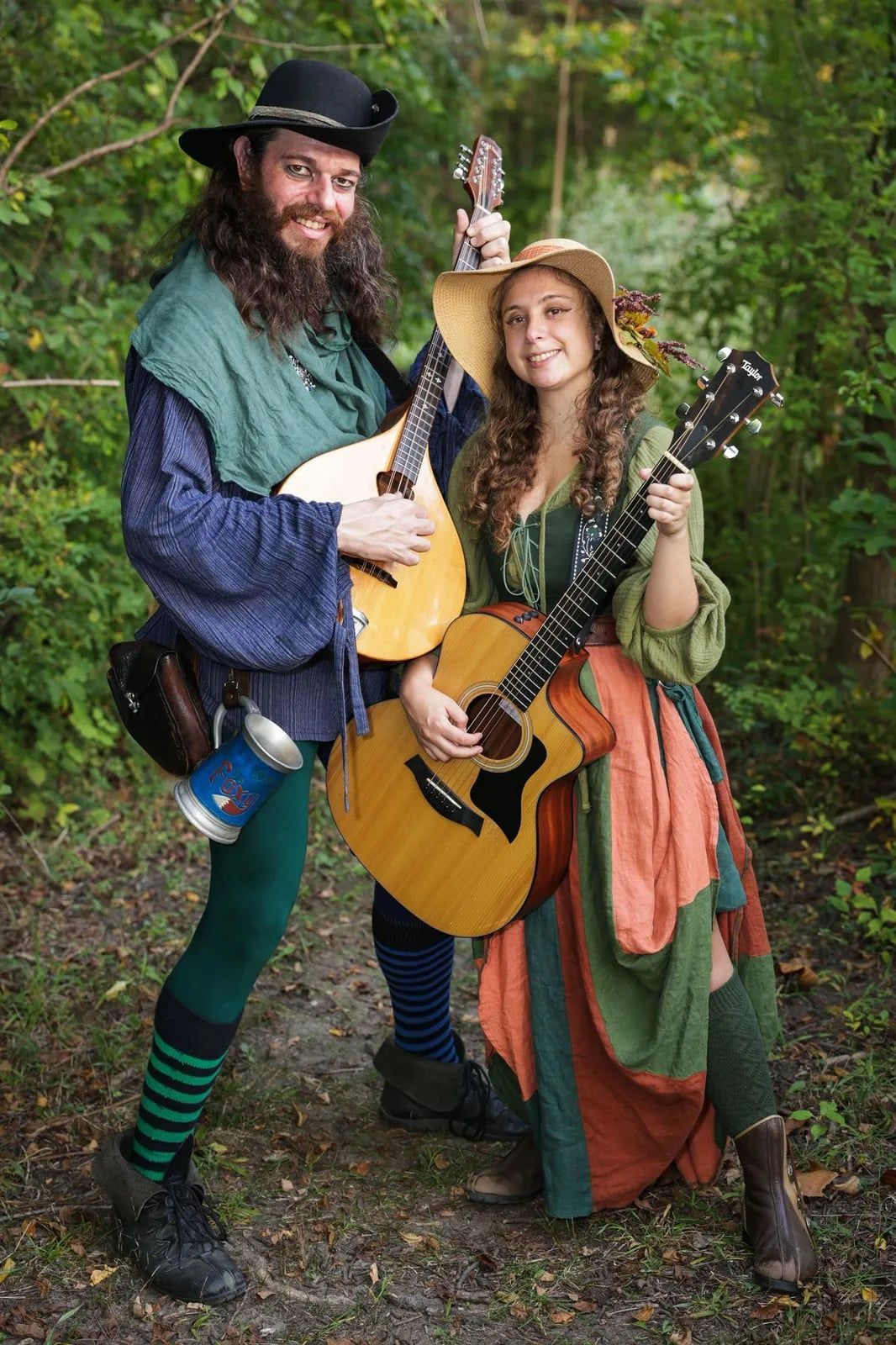 Man with mandolin and woman with guitar pose in wooded area, both in medieval attire.