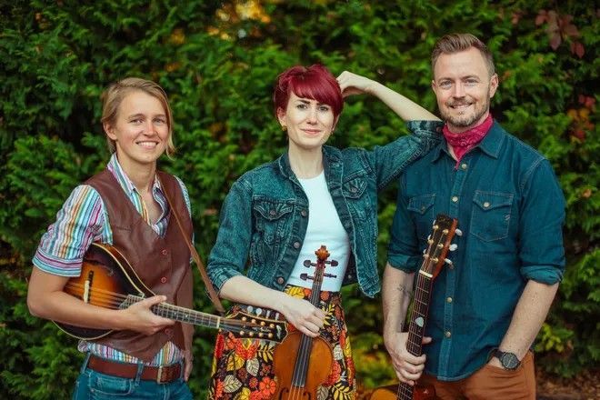 Three musicians posing outdoors with mandolin, violin, and guitar in front of greenery