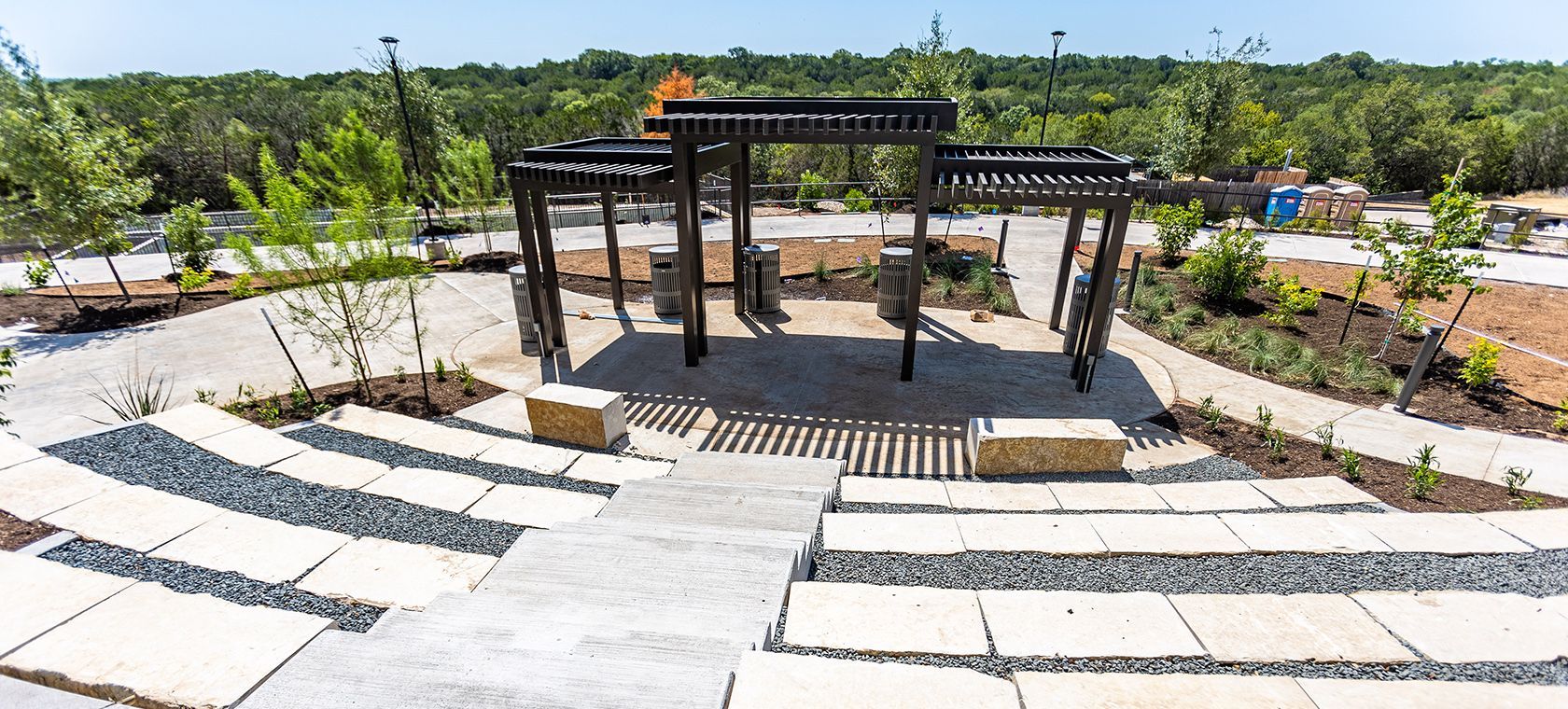 Outdoor amphitheater with black pergolas, white curved steps, and young trees in a landscaped park