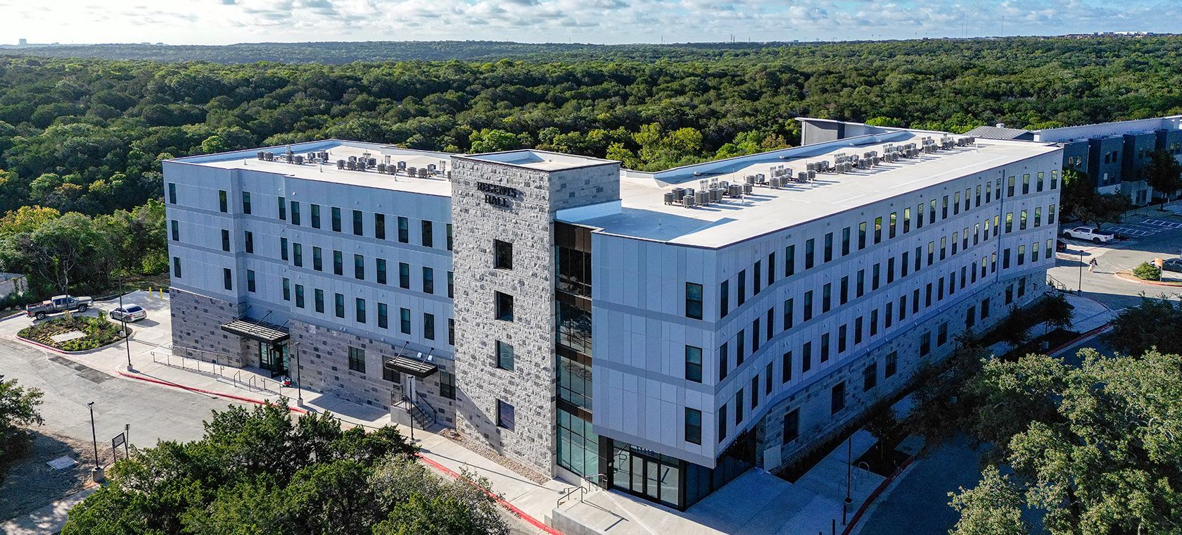 Aerial view of a modern white office building surrounded by trees and parking lots.