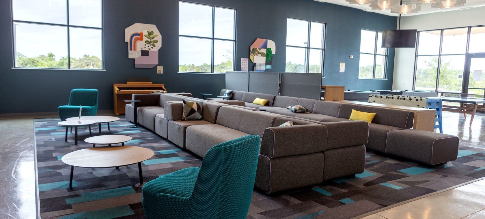 Modern lounge with gray sofas, teal chairs, round tables, and large windows in a spacious office lobby