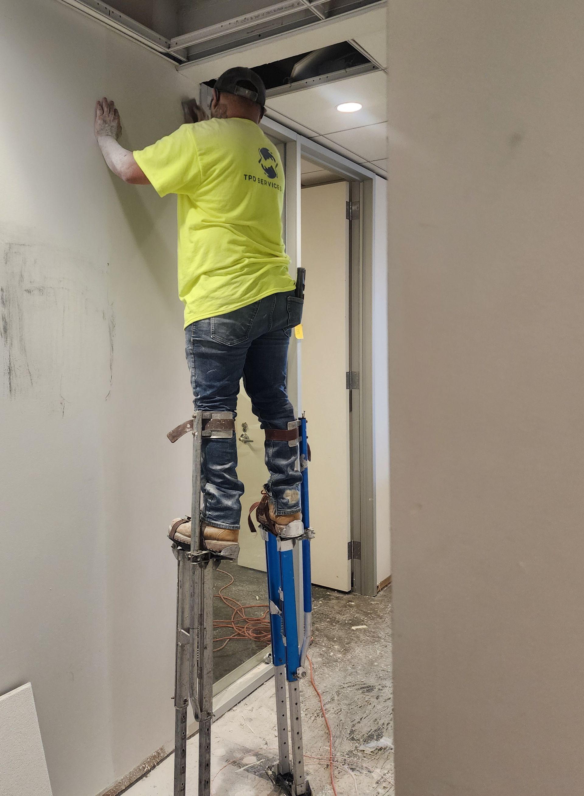 Worker in neon yellow shirt on a ladder painting a wall in a hallway.