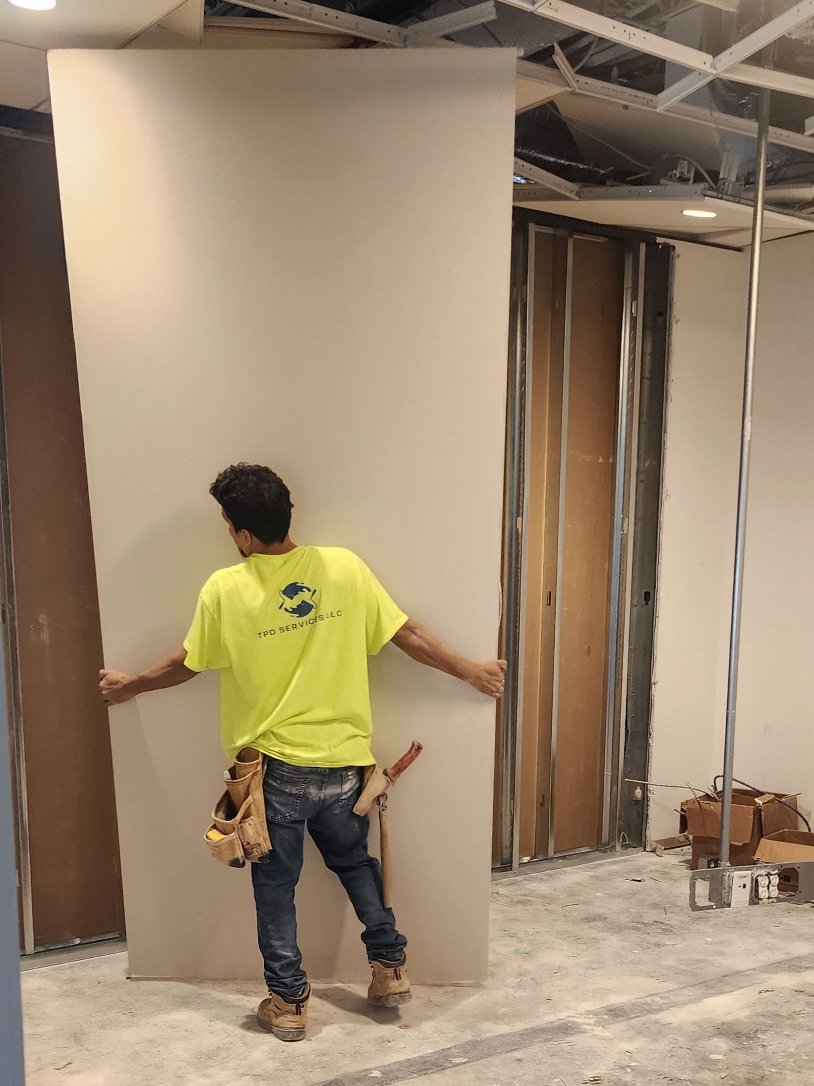 Person in a neon yellow shirt measuring a large wall in an unfinished interior space.