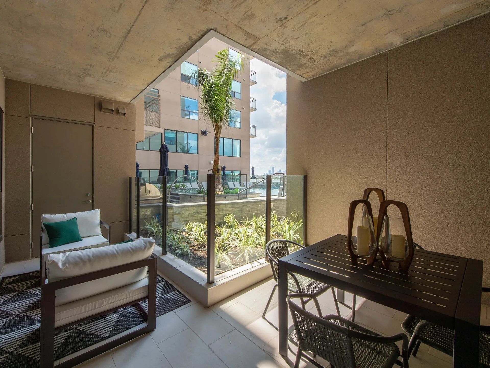Covered balcony with chair, dining table, and view of apartment building and greenery through glass railing.