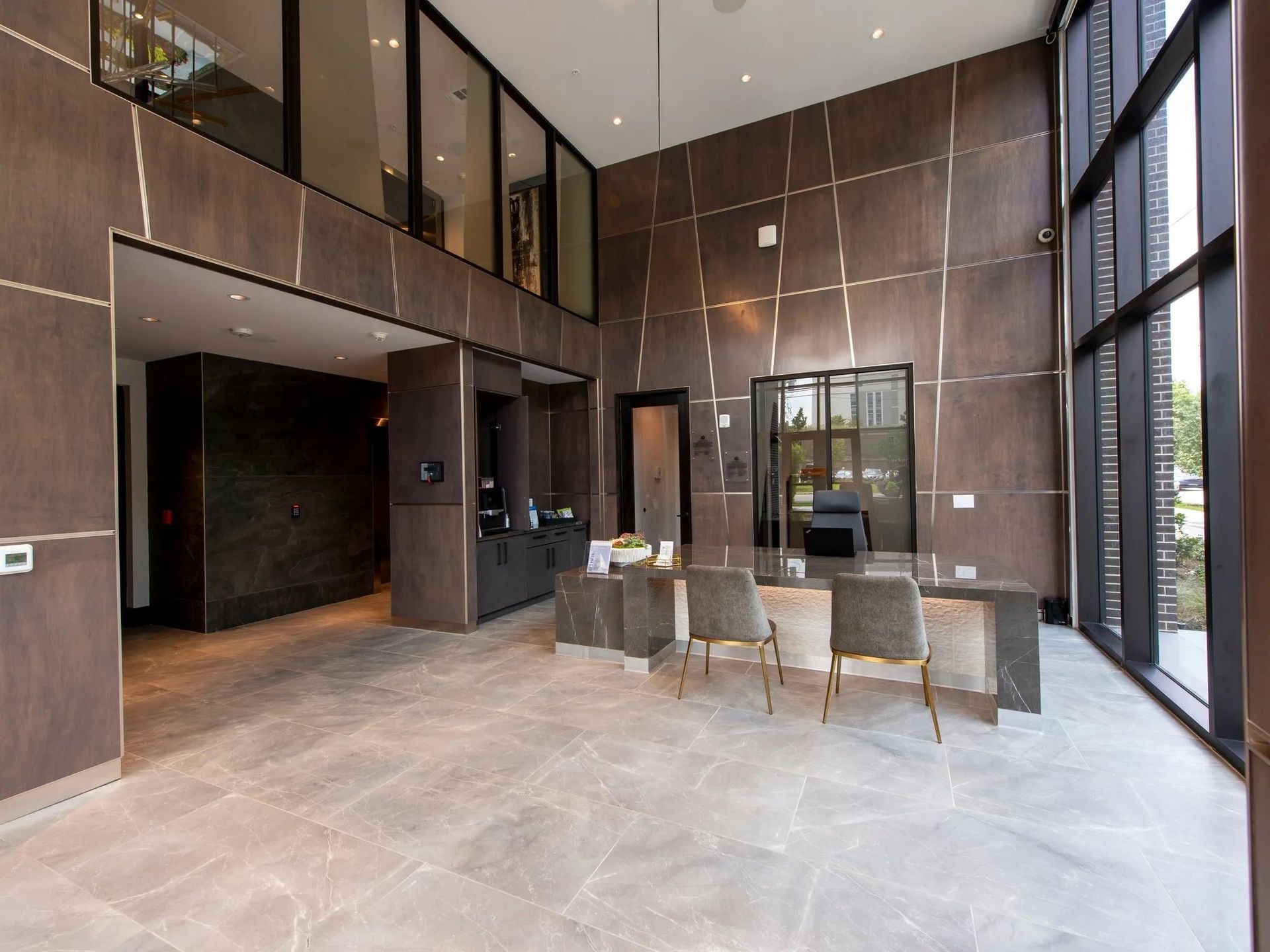 Modern lobby with tall windows, dark wood walls, and a glass table with chairs on a stone floor