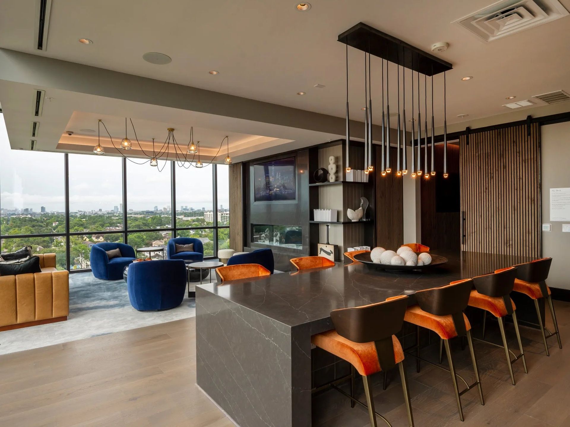 Modern hotel lounge with a dark kitchen island, orange stools, blue chairs, and city-view windows