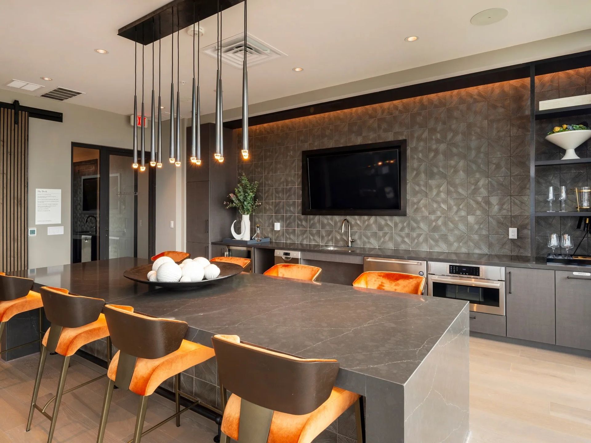 Modern kitchen with dark island, orange barstools, pendant lights, and wall-mounted TV