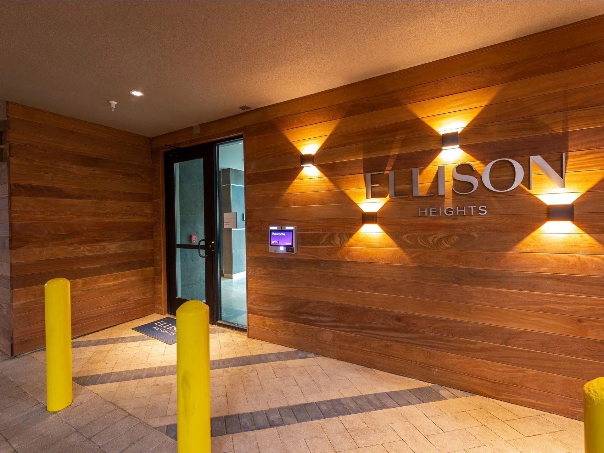 Lobby entrance with wood-paneled wall, illuminated ELISON RESIDENTS sign, and yellow bollards outside glass doors