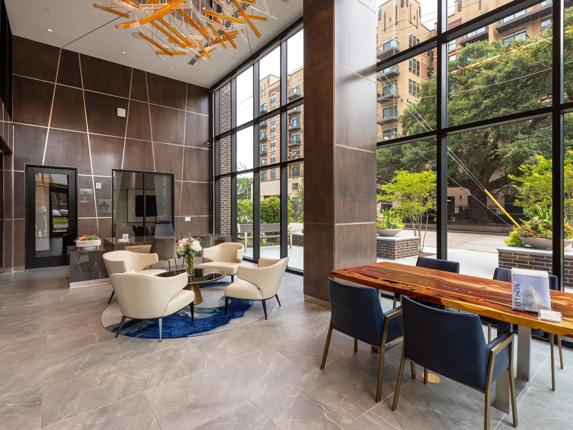 Modern hotel lobby with tall windows, beige seating, blue chairs, and a wooden table by the entrance.