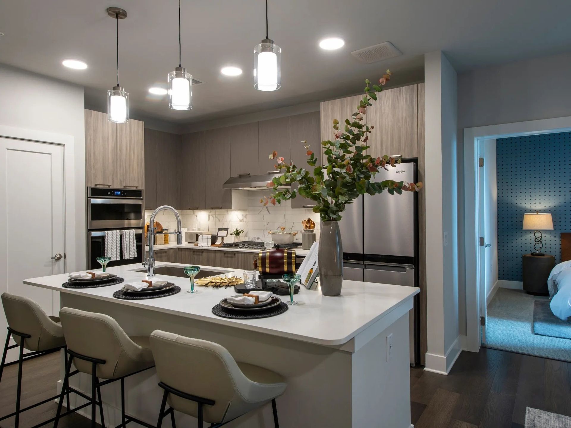 Modern kitchen with white island, pendant lights, and a dining setup, opening to a blue-lit bedroom.