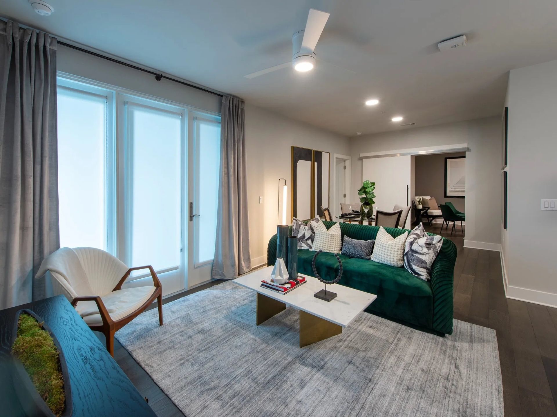 Bright modern living room with teal sofa, gray chairs, coffee table, rug, and large windows