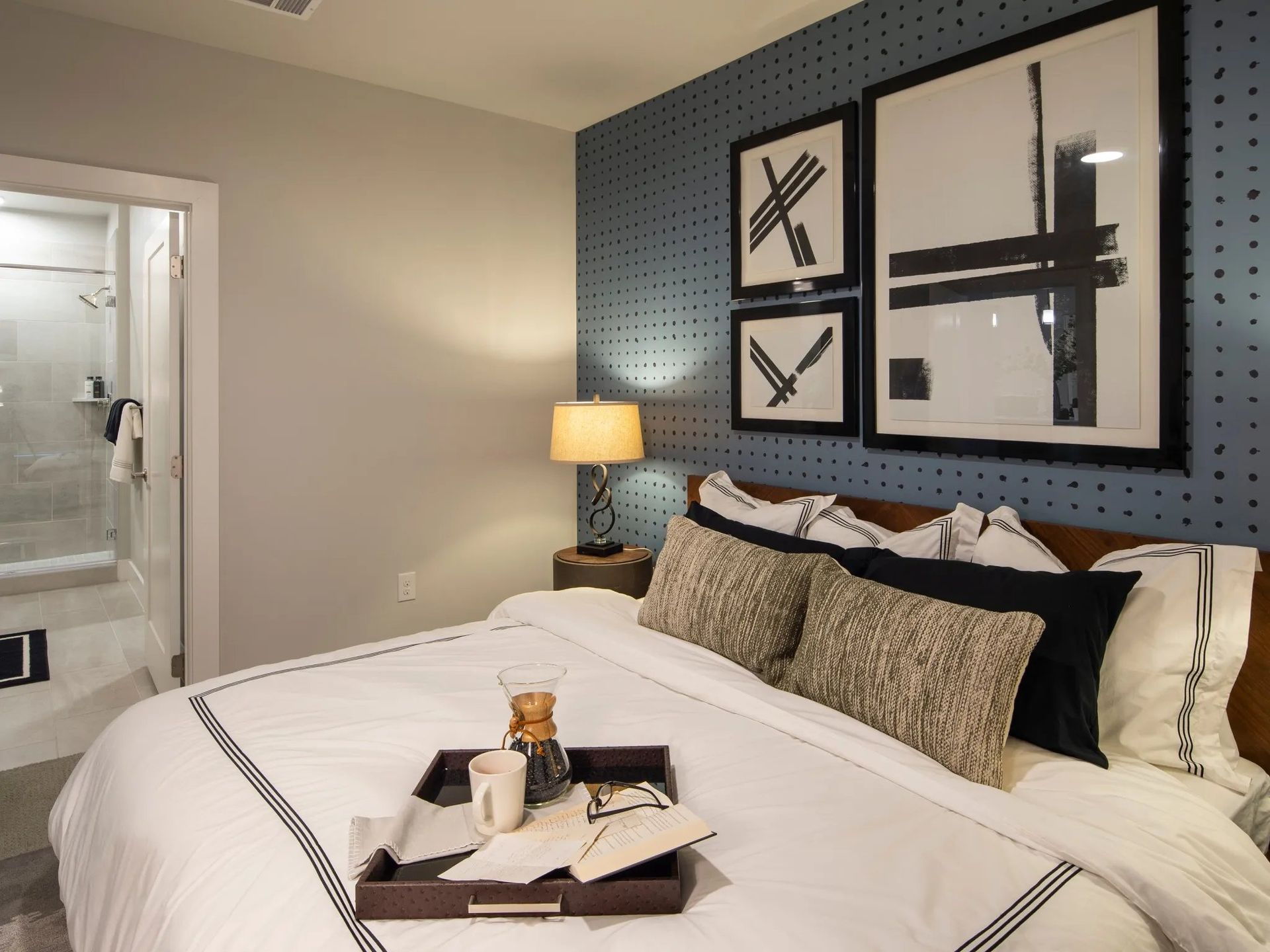 Neatly made hotel bedroom with white bed, blue accent wall, framed art, and lit bedside lamp.