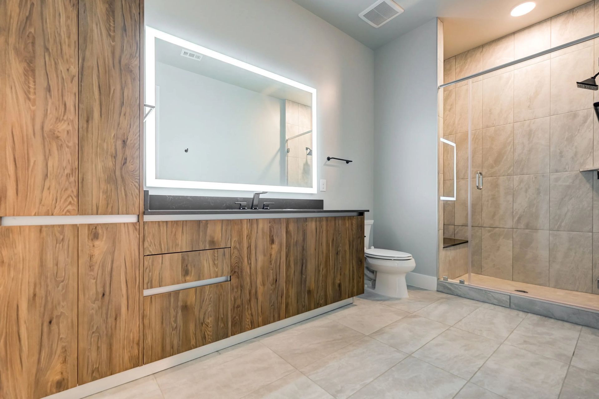 Modern bathroom with wood vanity, lit mirror, toilet, and glass shower