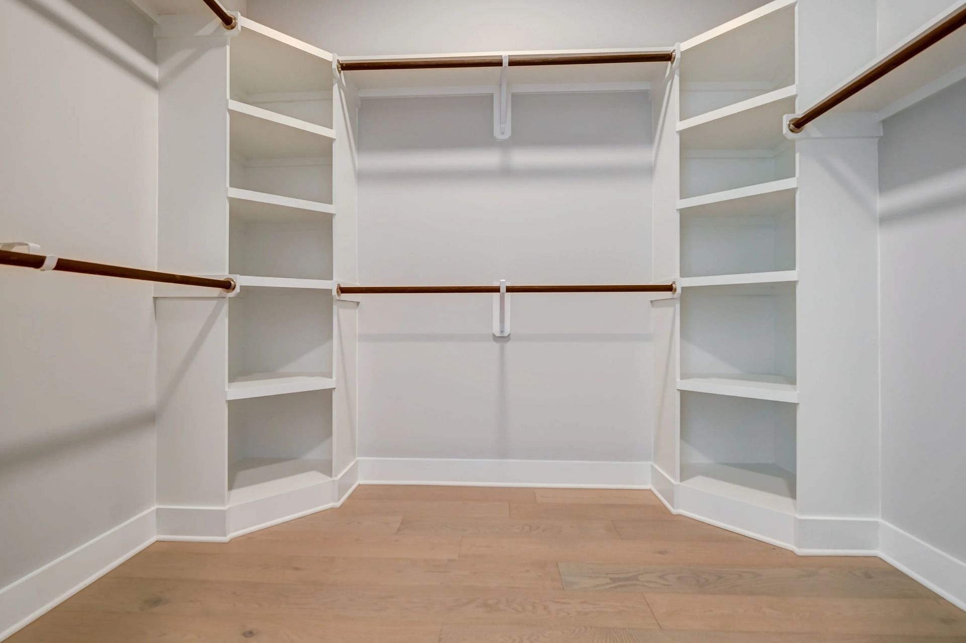 Empty walk-in closet with white shelving, hanging rods, and light wood flooring.