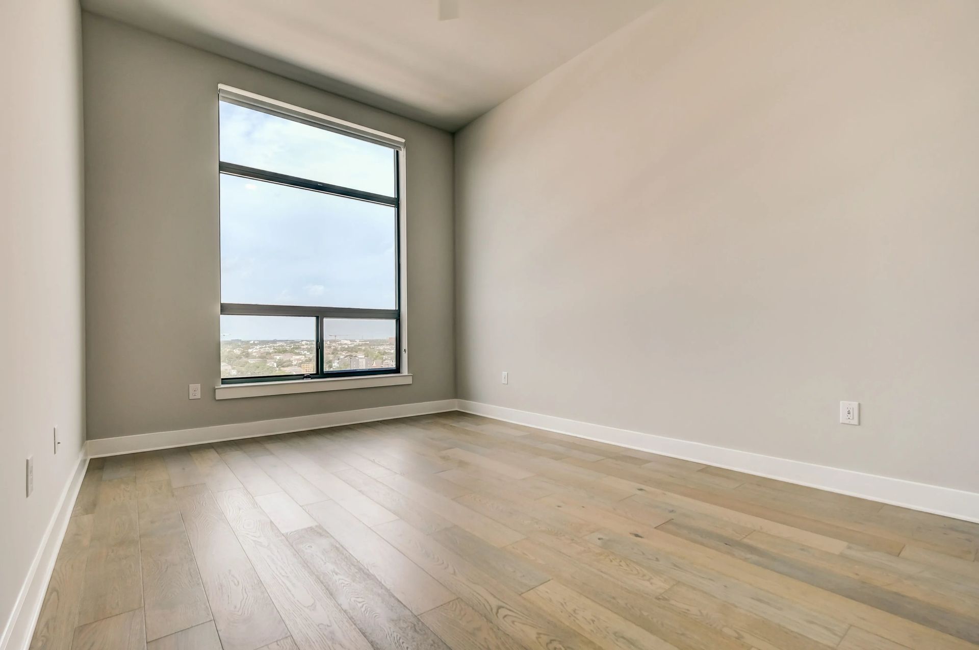 Empty modern room with light wood floors, large window, and white walls.