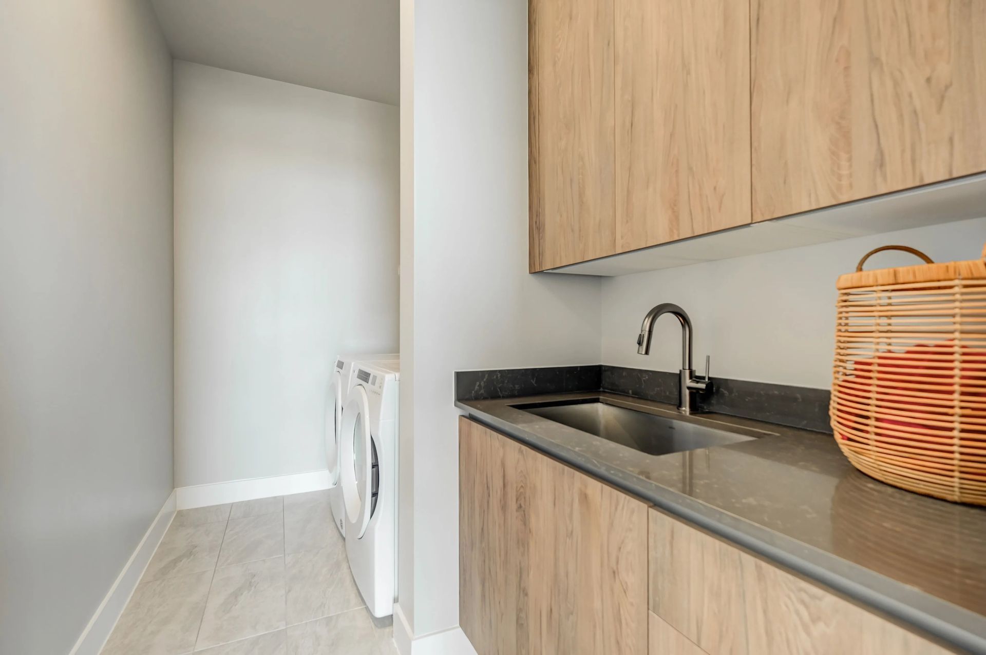 Compact kitchenette with wood cabinets, stainless sink, black countertop, and wicker basket in a narrow hallway.