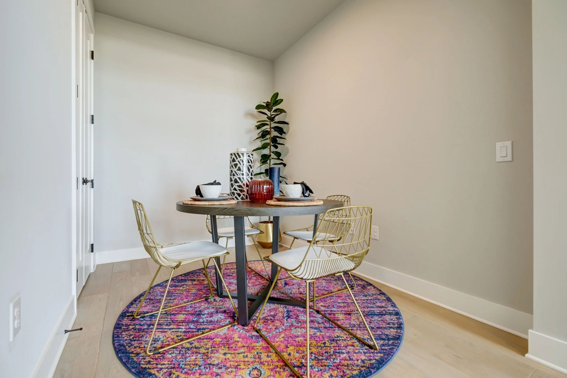 Small dining nook with round table, two chairs, colorful rug, and potted plant against white walls