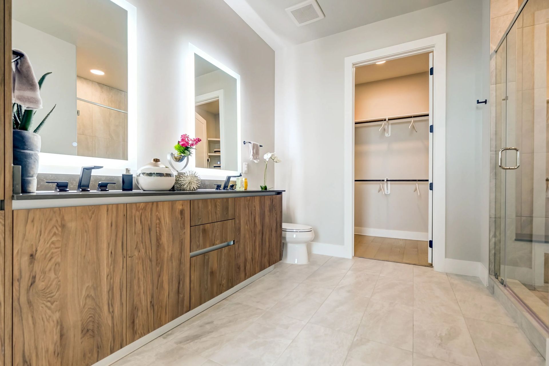 Modern bathroom with wood vanity, mirrors, glass shower, and open closet in a bright neutral interior