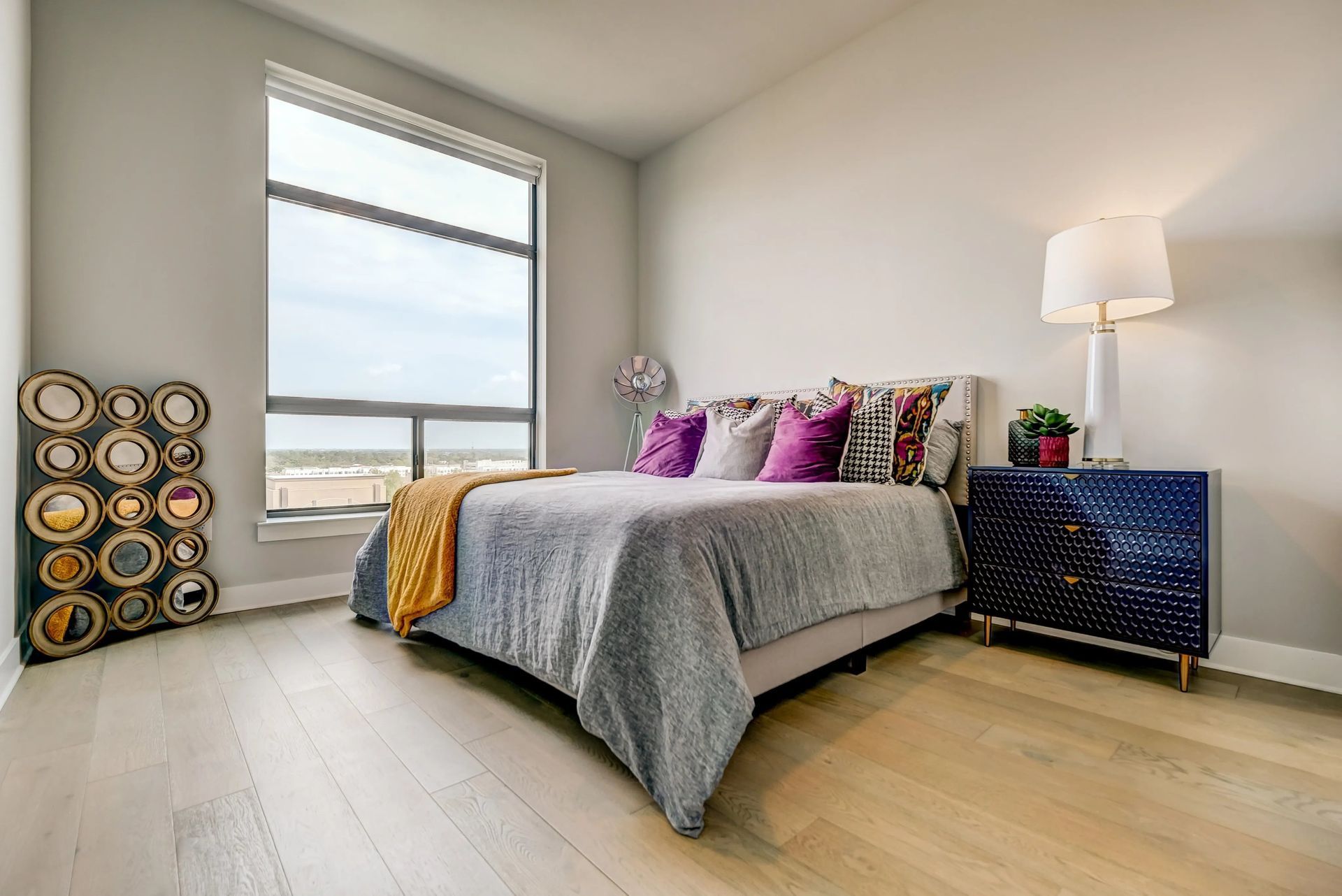 Bright bedroom with a gray bed, colorful pillows, large window, and blue nightstand with lamp.