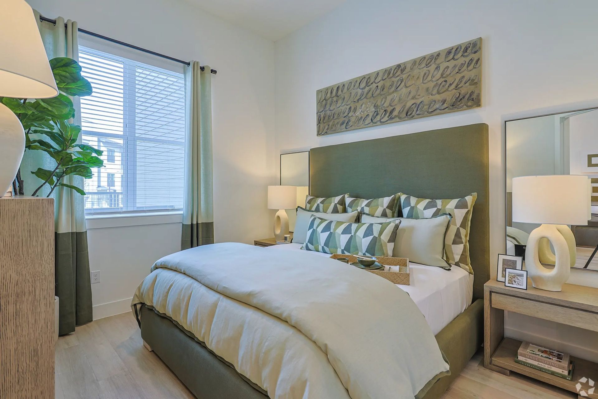 Bright modern bedroom with large bed, window blinds, bedside lamp, and vanity mirror