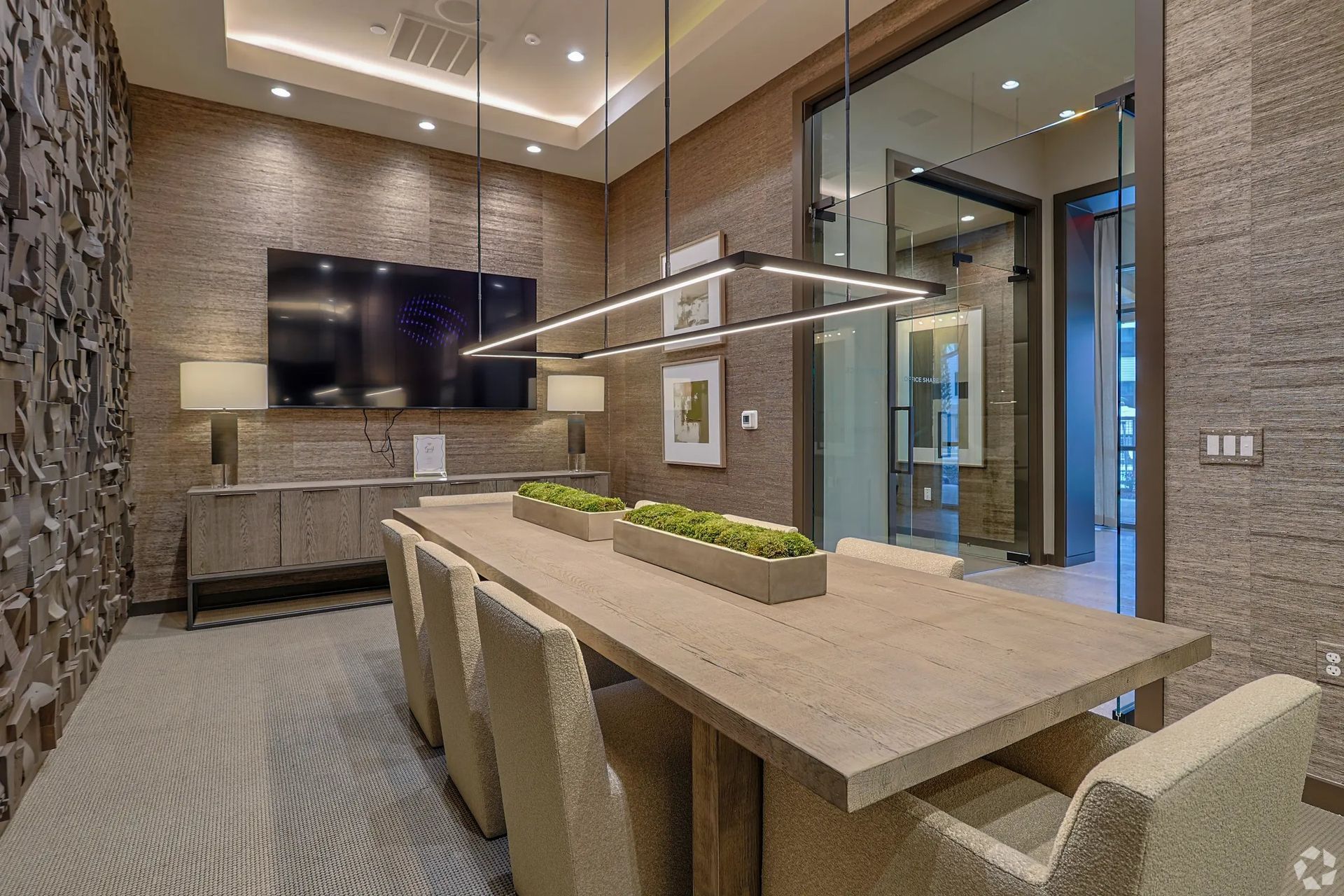 Modern conference room with a long table, beige chairs, wall-mounted TV, and glass doors