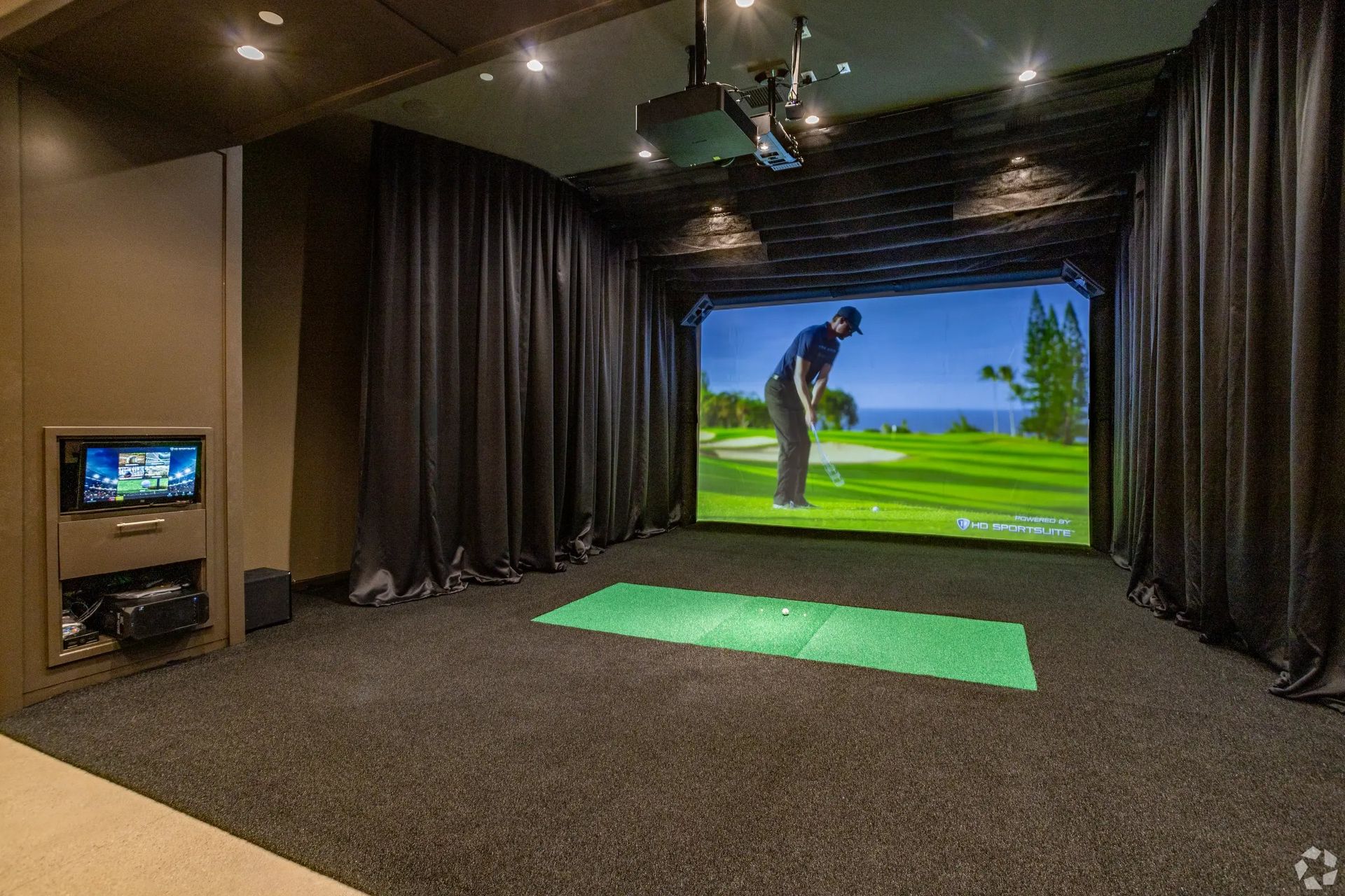 Indoor golf simulator room with a large screen, green hitting mat, and curtains around the bay.
